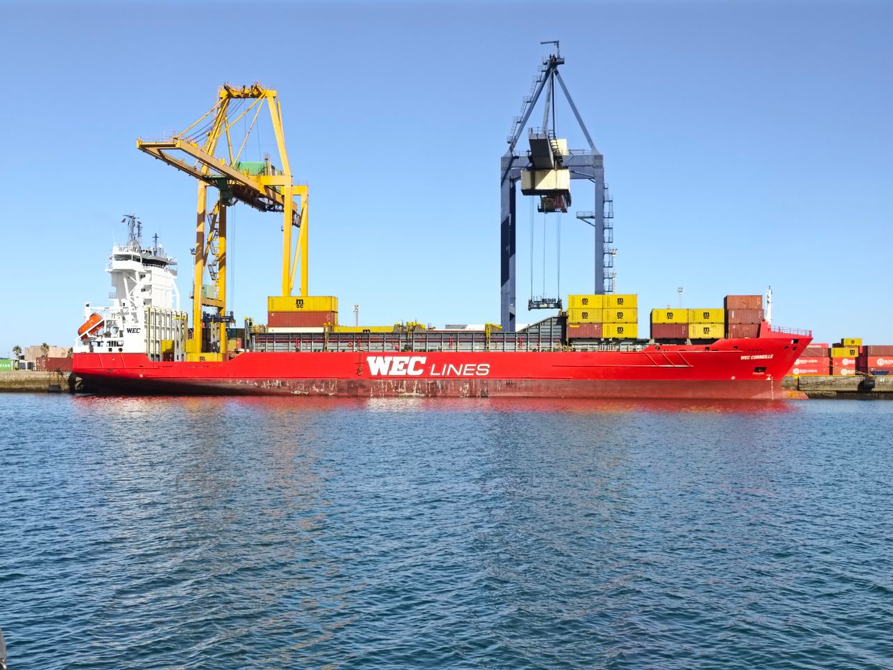 A red cargo ship, WEC Lines, docked at the busy port in Cádiz, Spain, under clear skies.